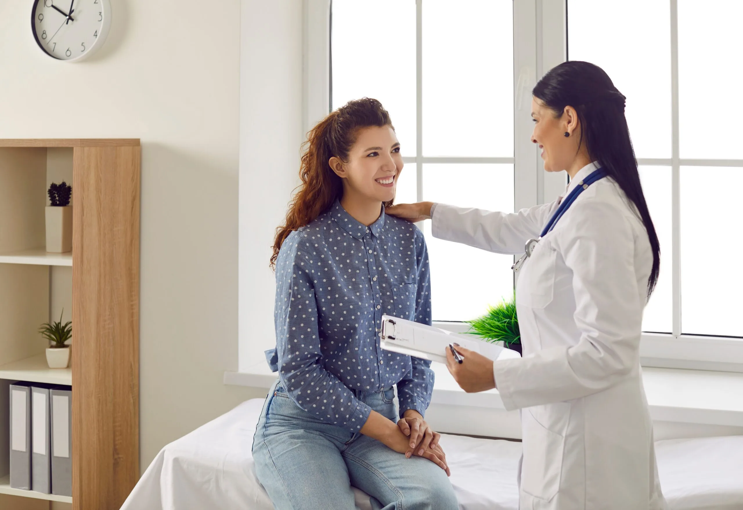 Female doctor in white coat touches patient's shoulder reassuringly while holding clipboard in bright office