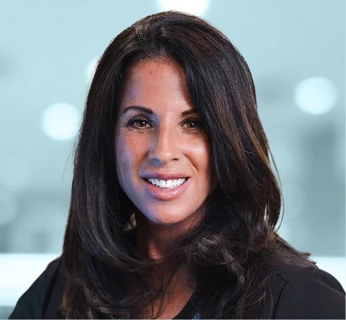 Woman with long dark hair smiling at camera against blurred light background