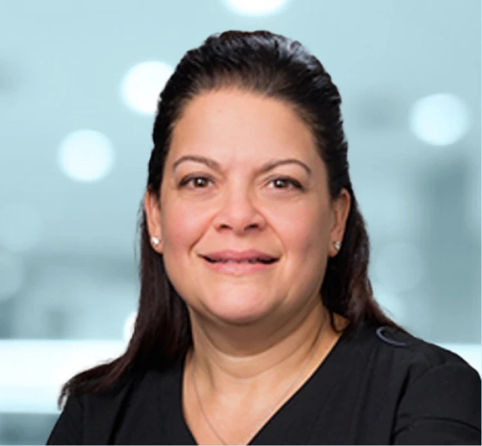 Woman with long dark hair pulled back smiling at camera against blurred light background