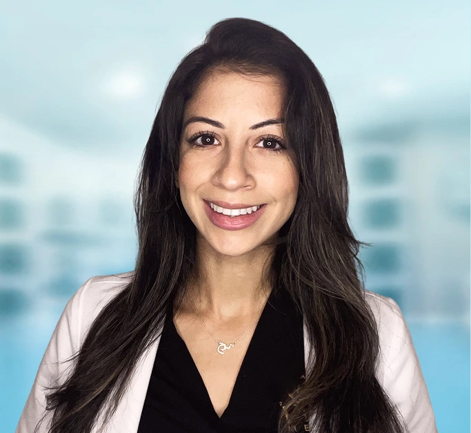 Woman with long dark hair and pendant necklace smiling at camera against blurred office background