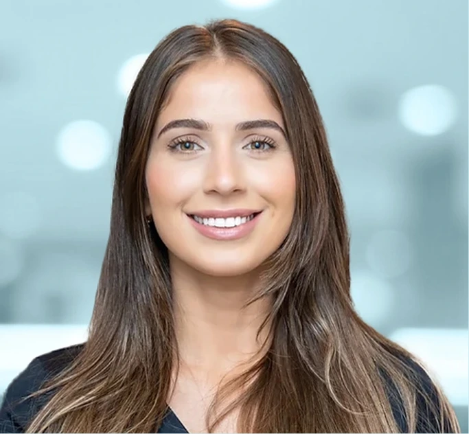 Young woman with long brown hair smiling at camera against blurred light background
