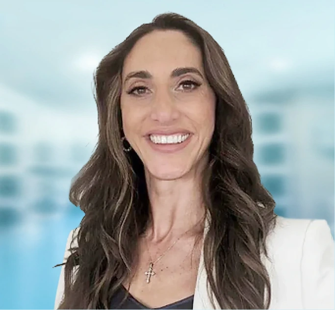Woman with long dark hair wearing white blazer smiling at camera against blurred light background