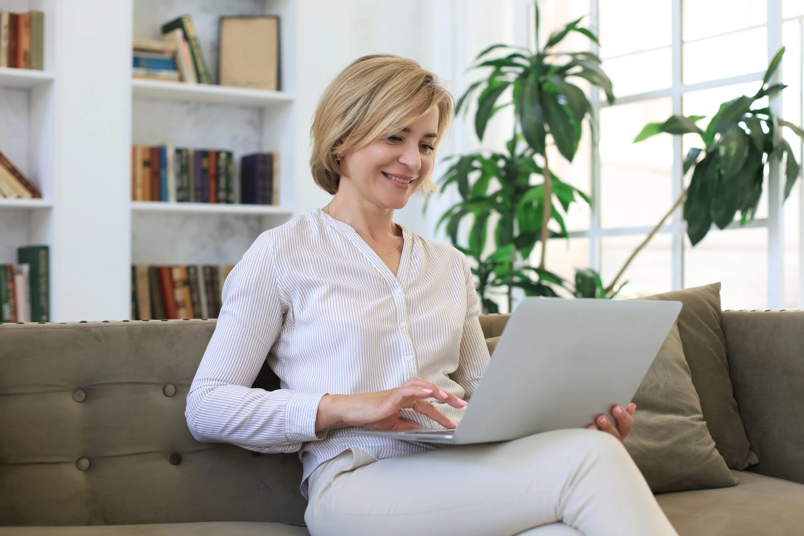 Middle-aged woman smiling while using laptop on sofa in bright living room with bookshelves and plants