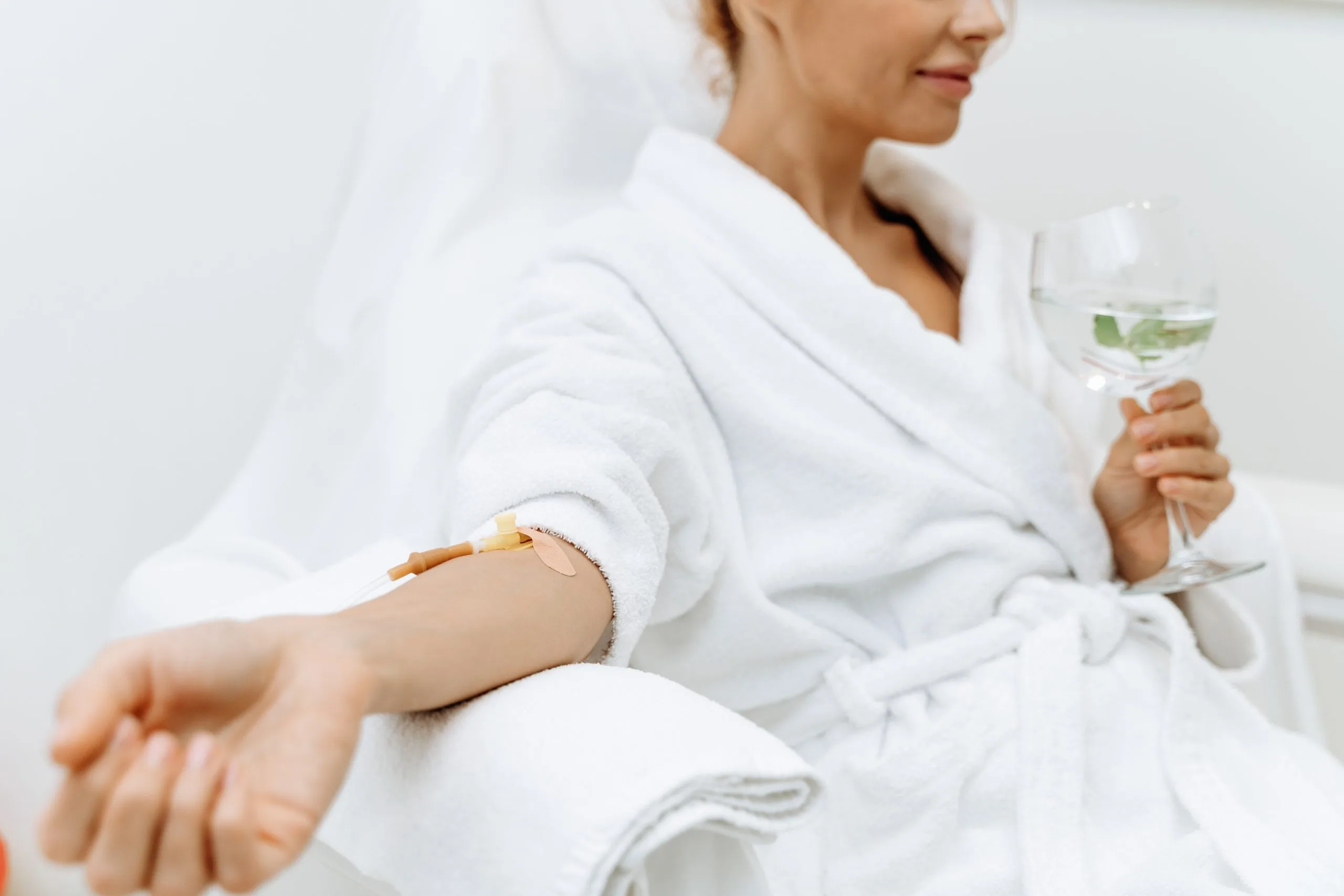 Woman in white robe holding water glass with bandage on arm after treatment