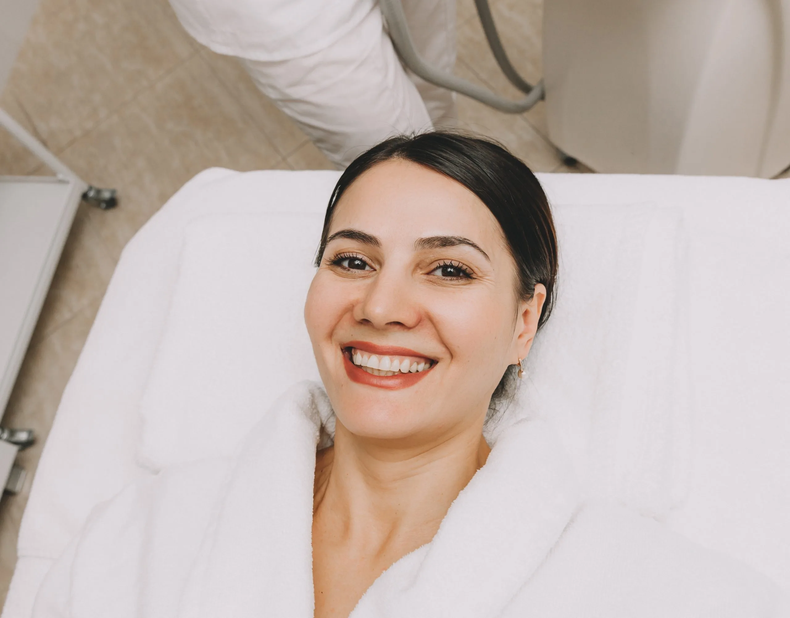 Woman smiling on white treatment bed wearing white robe in aesthetic clinic