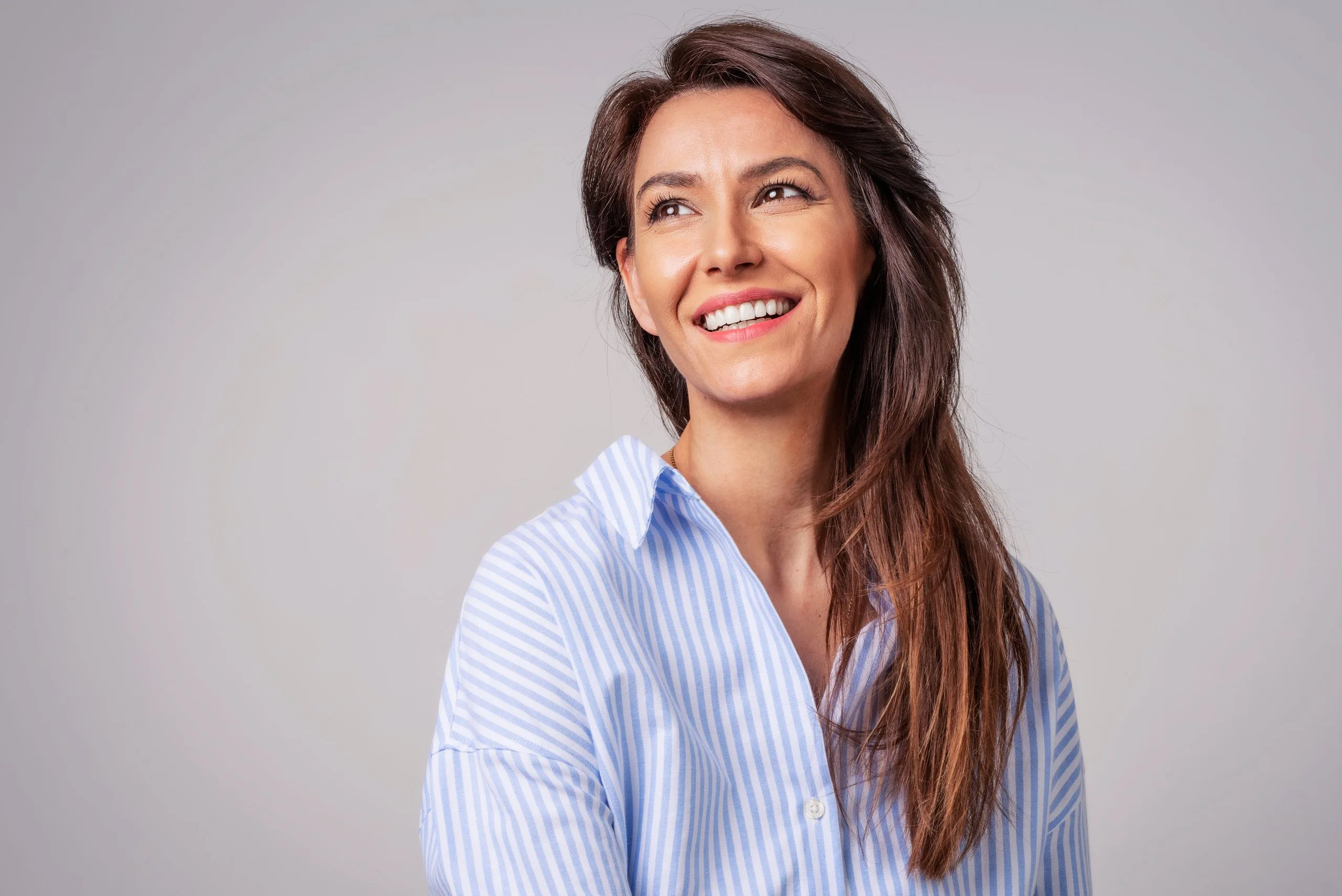 Woman smiling at camera wearing light blue striped shirt against gray background