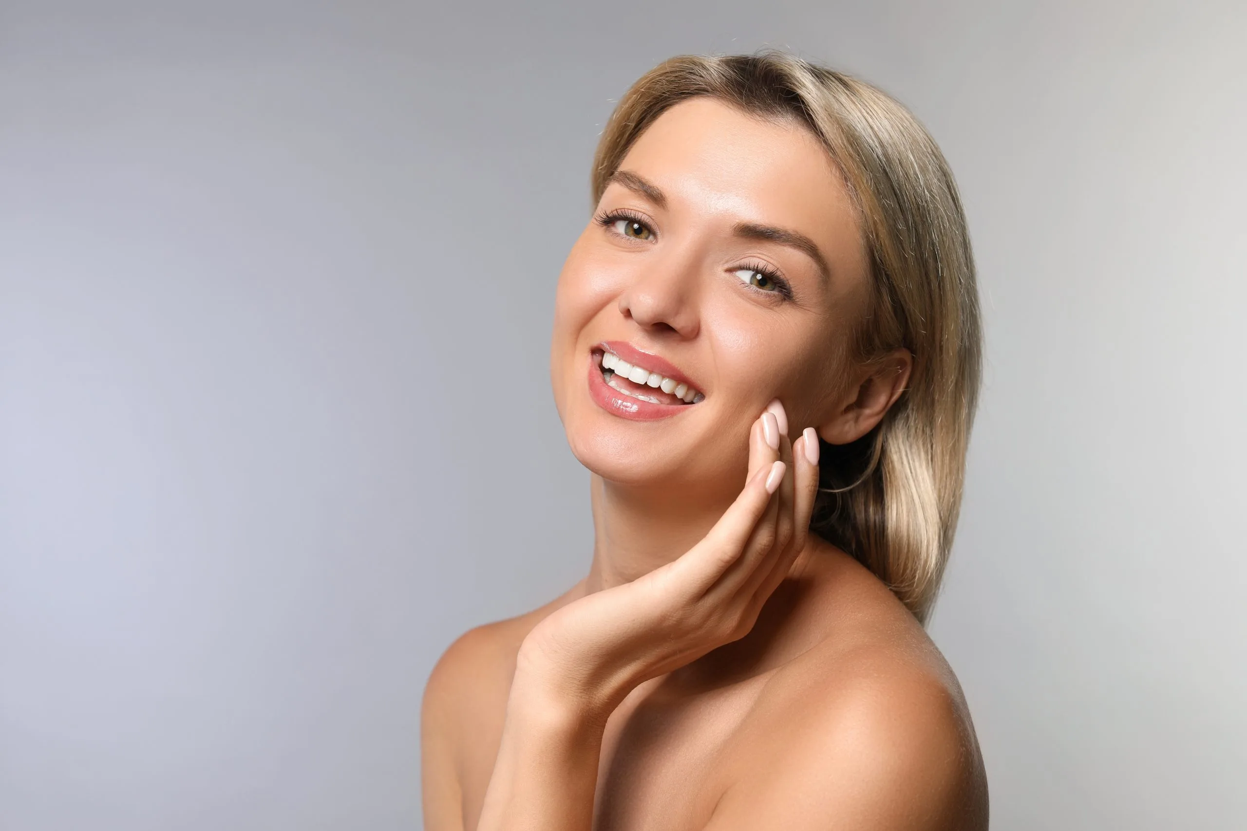 Smiling woman with blonde hair touching her cheek and jawline against a gray background