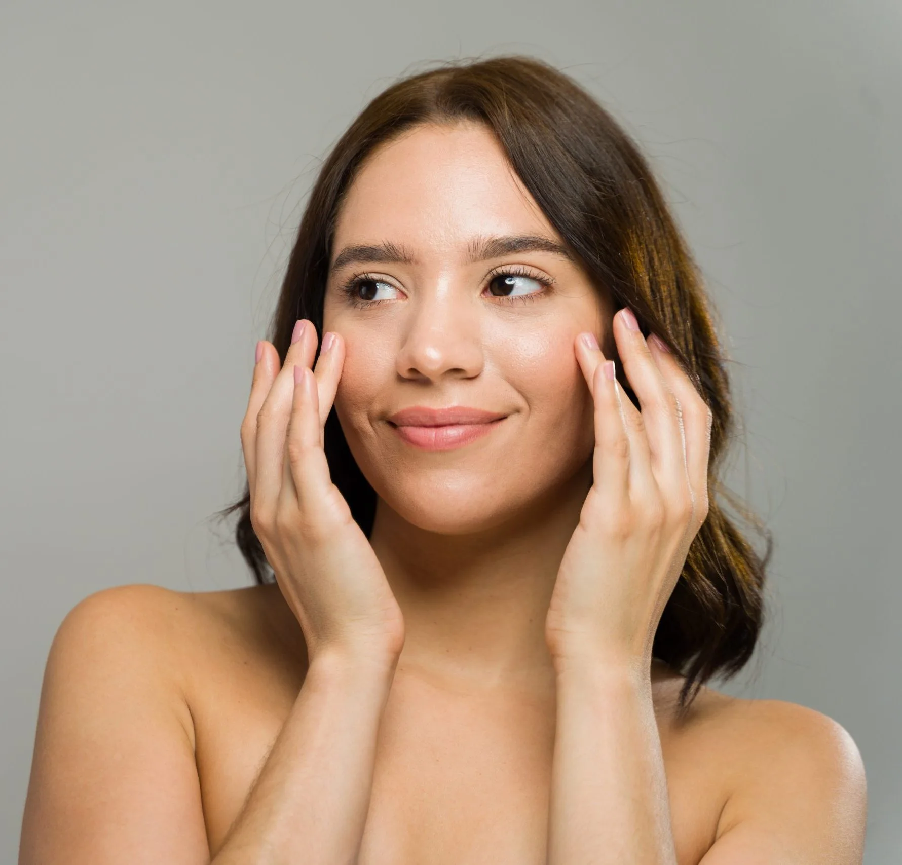 Woman touching her face with both hands, smiling at camera against gray background