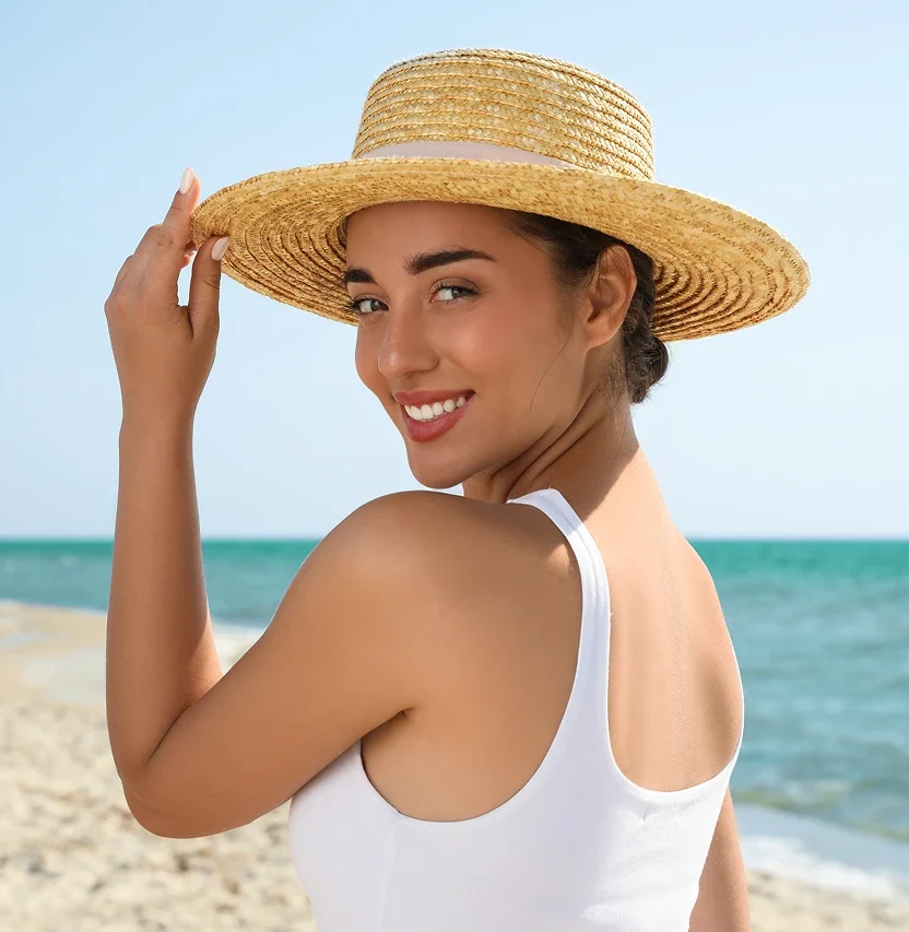 Woman in white tank top and straw hat smiling at beach with ocean behind her