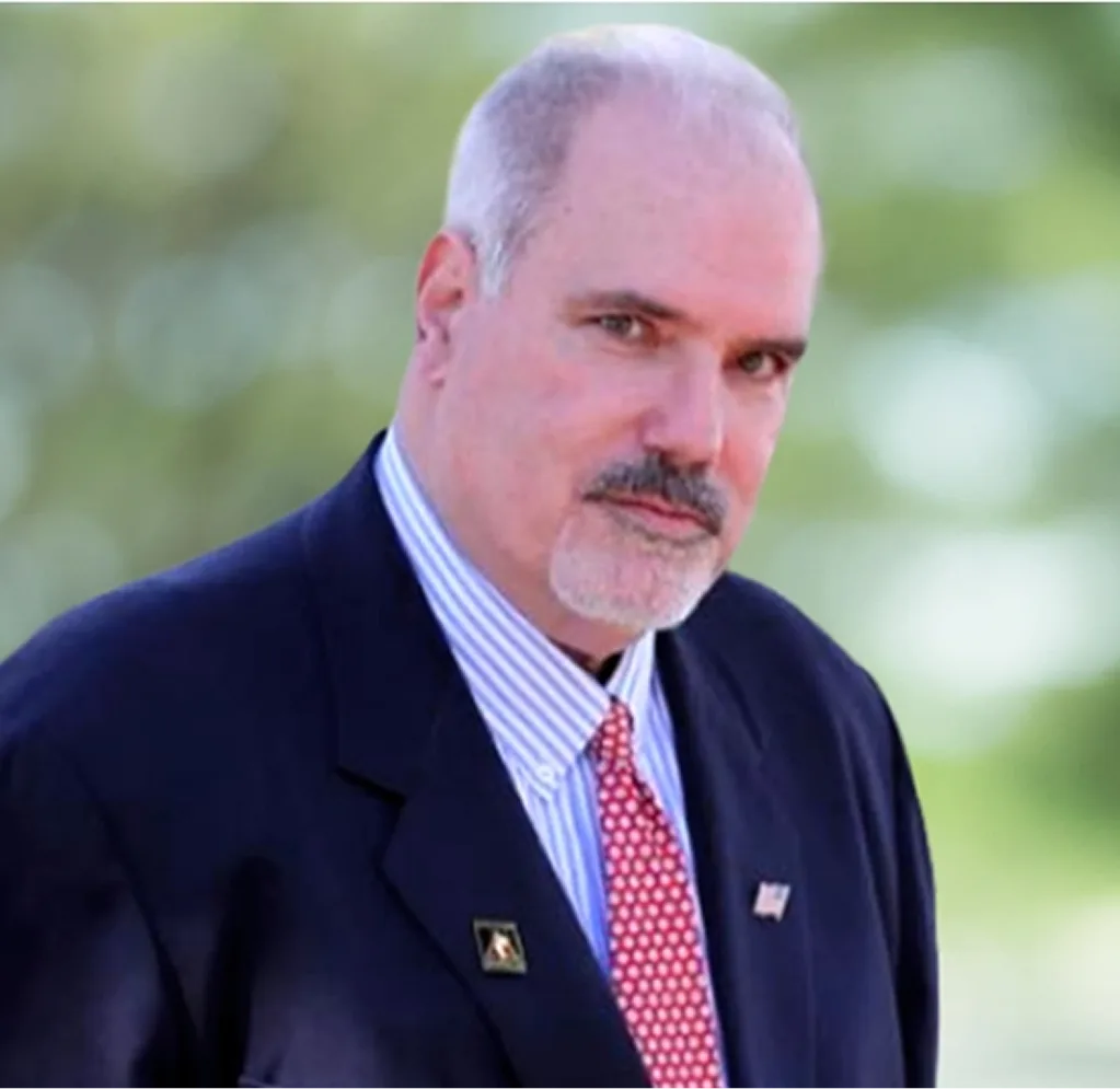 Professional headshot of man with mustache wearing dark blazer and red patterned tie against blurred green background