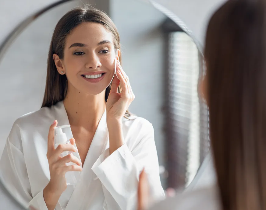 Woman in white coat applying skincare product to her face while looking in mirror