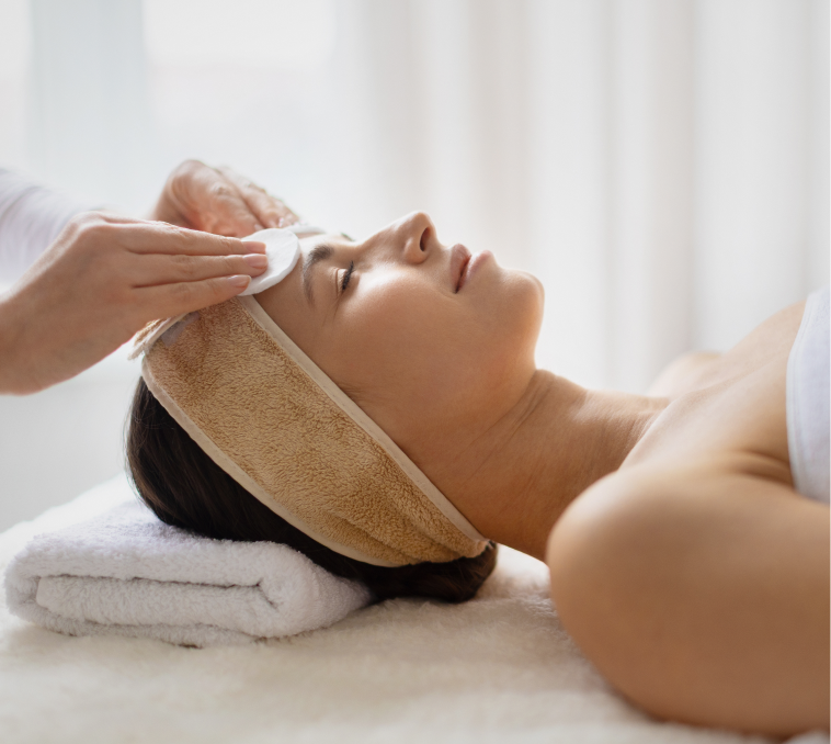 Aesthetician applying facial treatment to relaxed patient lying on spa bed with towel headwrap
