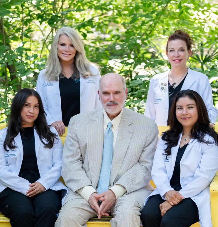 Five medical professionals in white coats posing together outdoors against green foliage