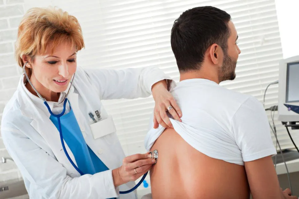 A female doctor listens to a male patient's back with a stethoscope during a physical exam.