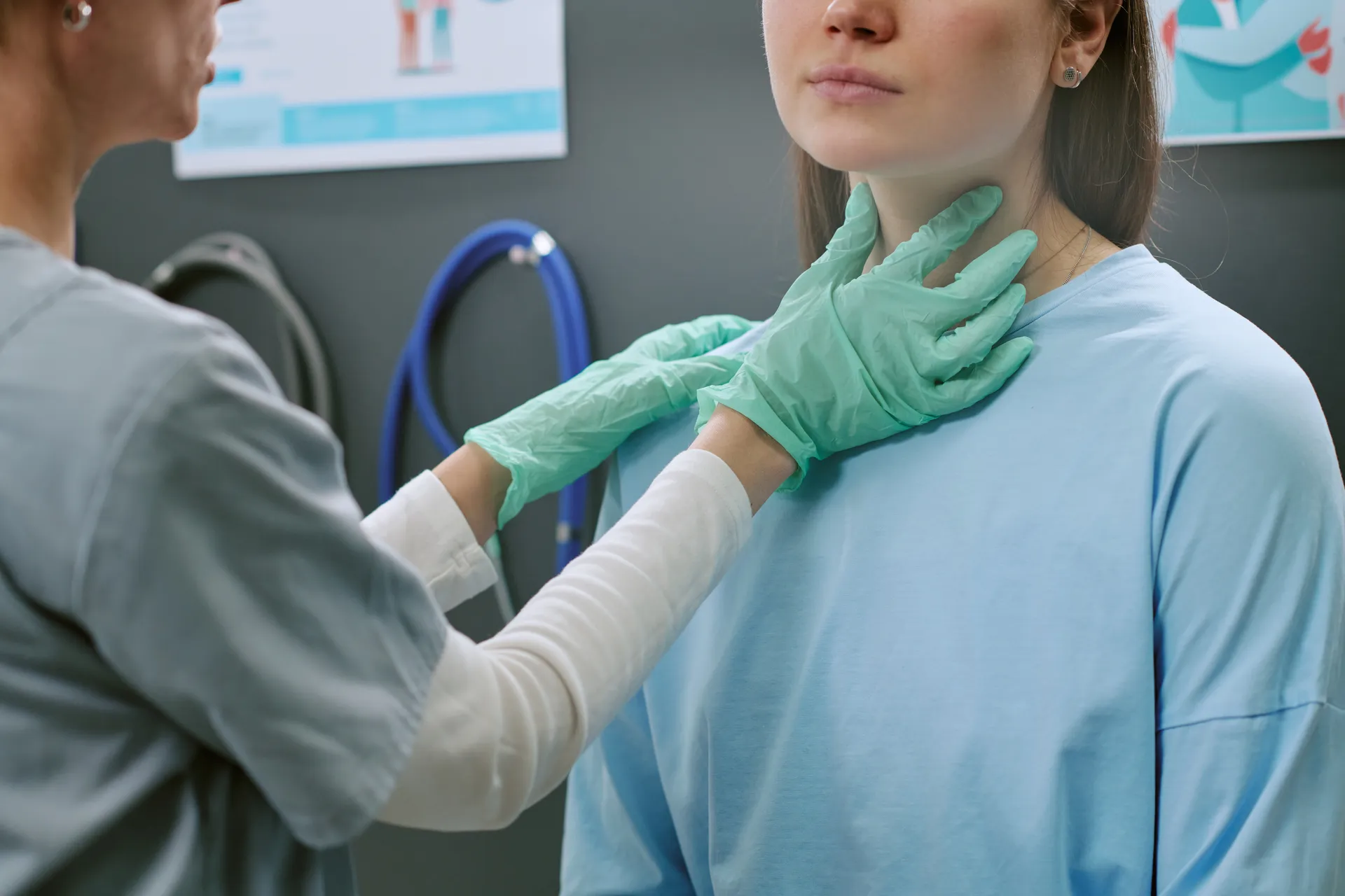 A gloved clinician palpates a female patient's thyroid gland during a neck examination in a clinical exam room.
