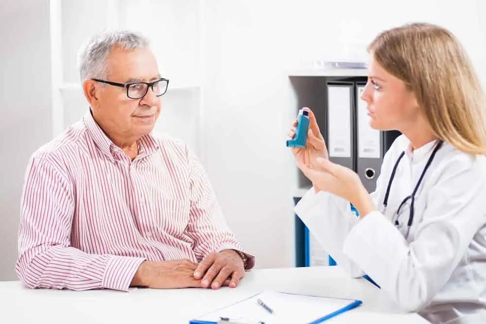 A female doctor explains the use of an asthma inhaler to an elderly male patient during a consultation.