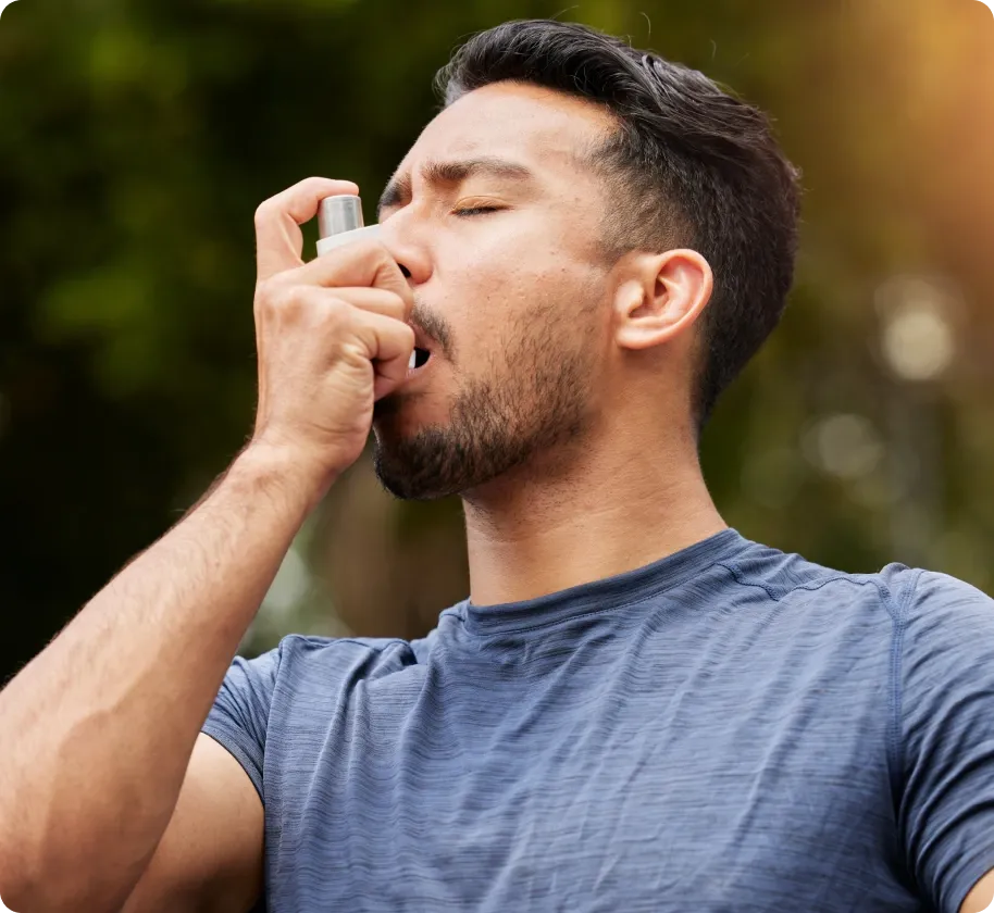 A man outdoors uses an asthma inhaler with eyes closed, managing a respiratory condition.