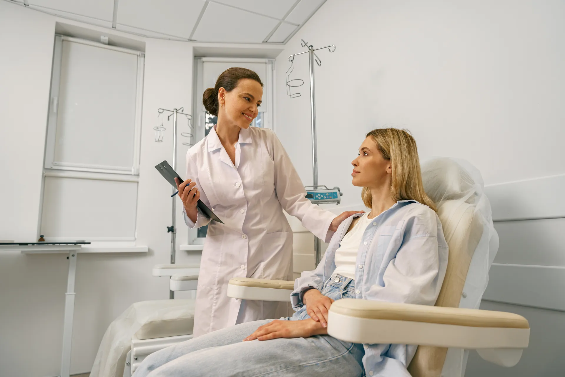 A smiling female doctor in a white coat consults with a seated patient in an infusion treatment room.