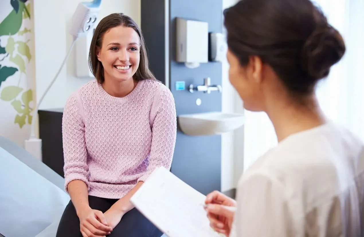 A smiling female patient in a pink sweater consults with a female provider holding a clipboard in a medical exam room.