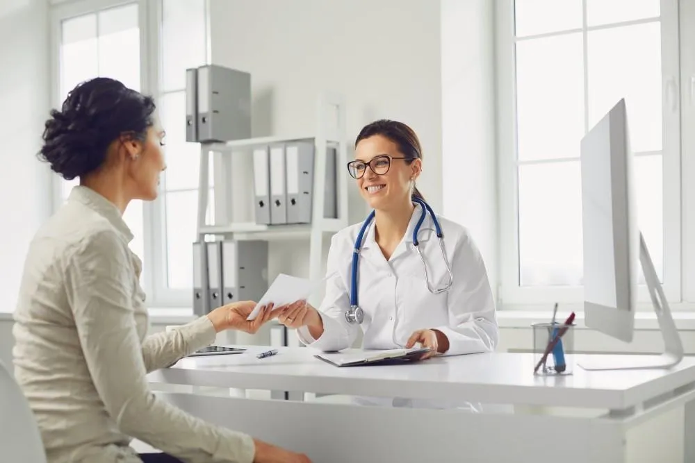 A smiling female doctor in a white coat hands a document to a patient during a consultation in a bright office.