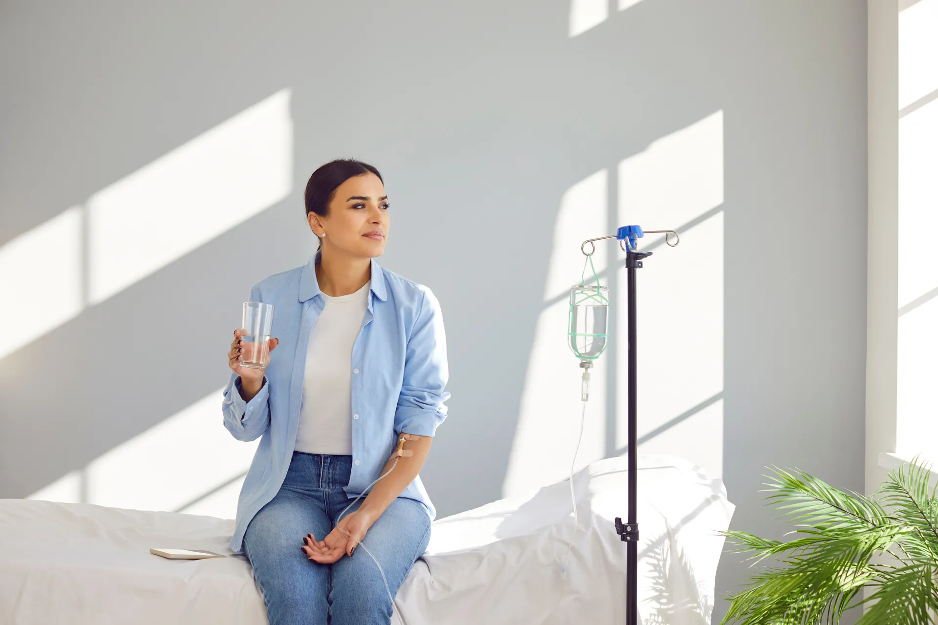A woman sits on a treatment bed holding a glass of water while receiving IV therapy in a bright, sunlit room.