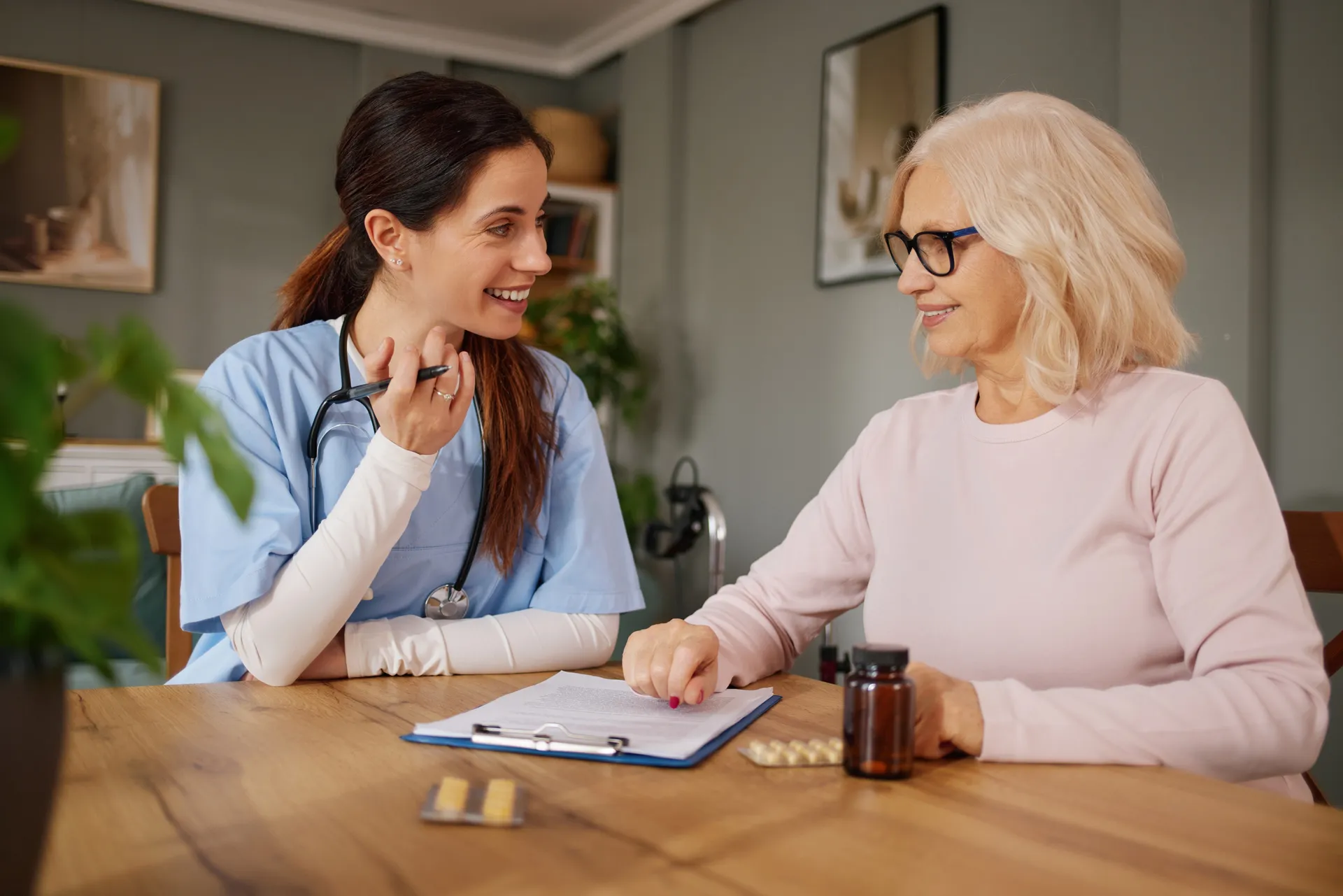 A smiling nurse practitioner reviews a medication management plan with an older female patient at a table with pill bottles nearby.