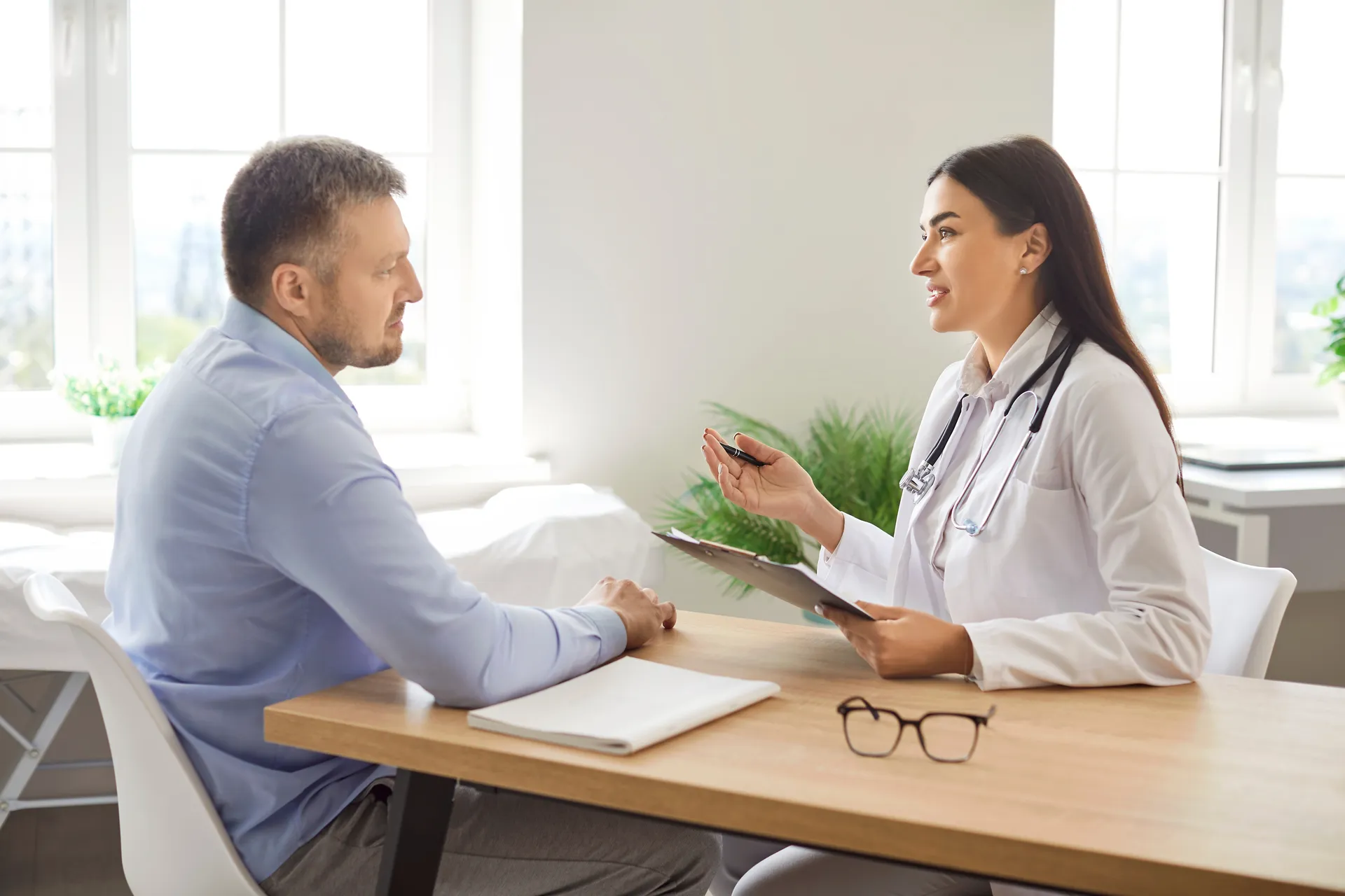 A primary care doctor with a clipboard talks a patient through a specialist referral at a desk.