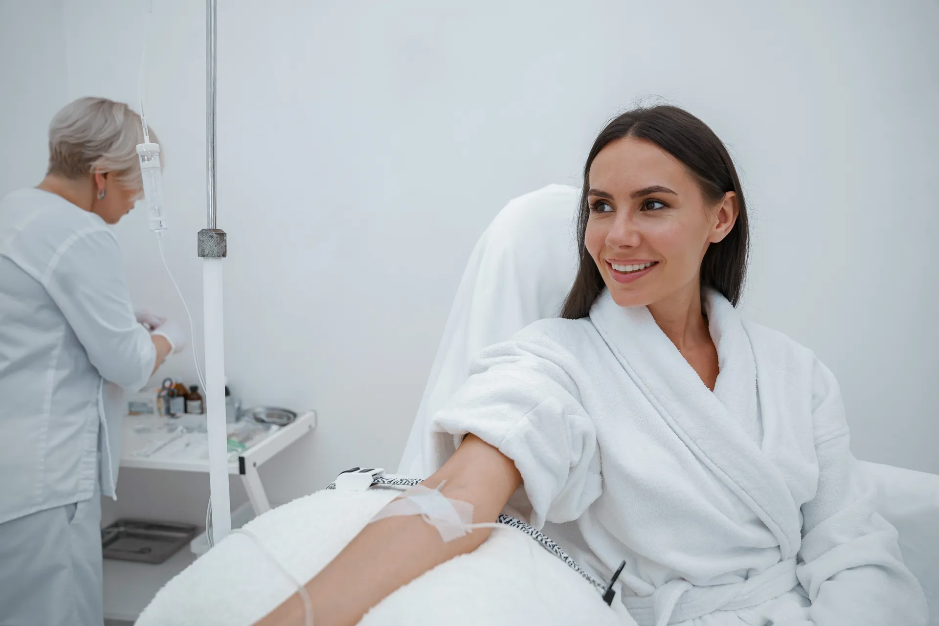A smiling woman in a white robe receives an IV infusion while a provider prepares supplies in the background.