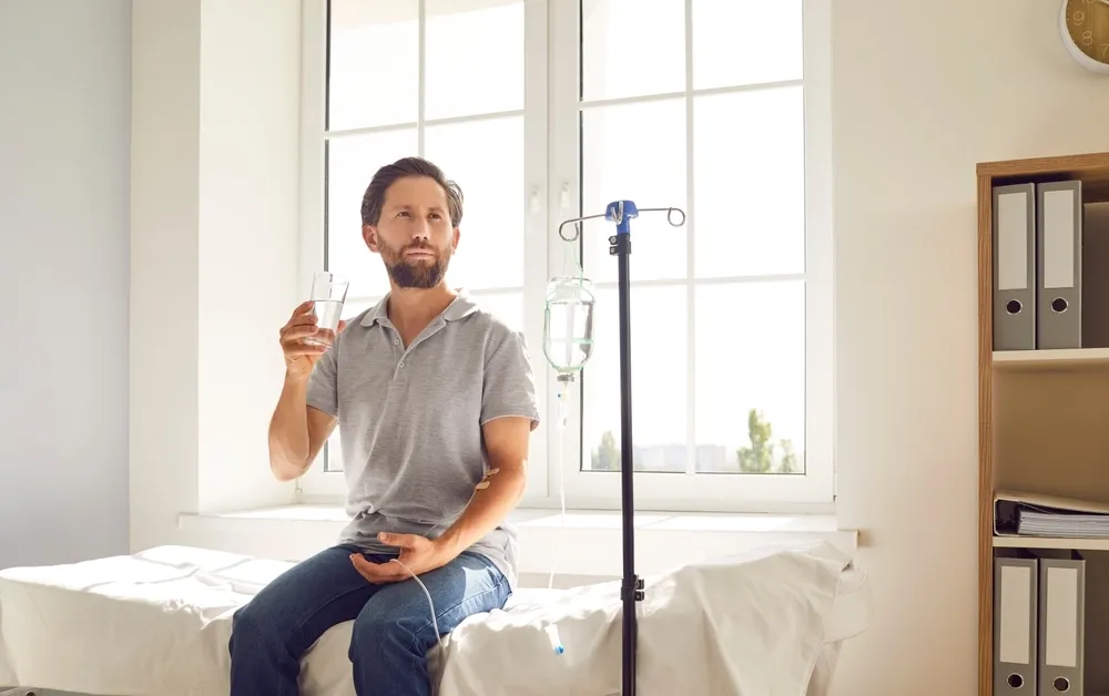 A man sits on a treatment bed receiving an IV infusion while drinking water in a bright clinical room.