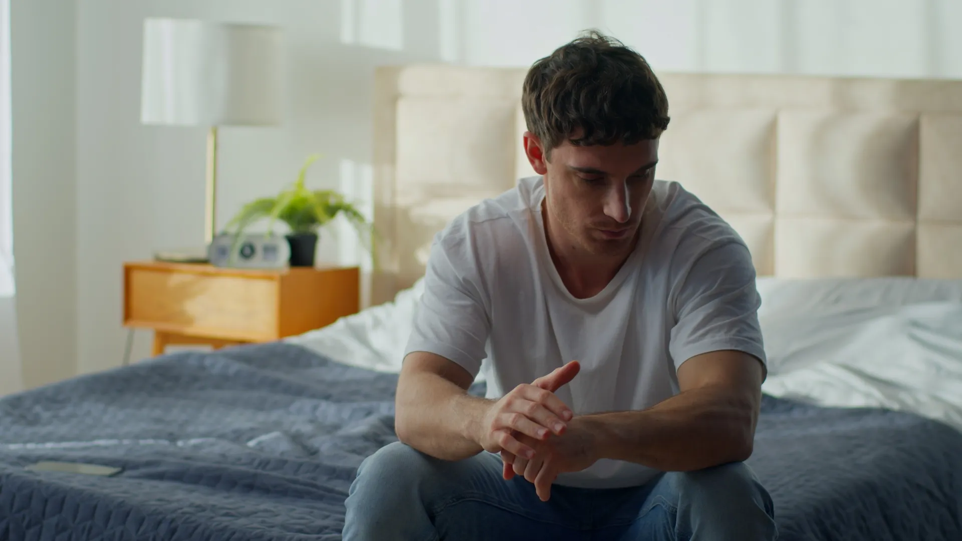 A pensive man sits on the edge of his bed with hands clasped, illustrating signs of low testosterone in men.