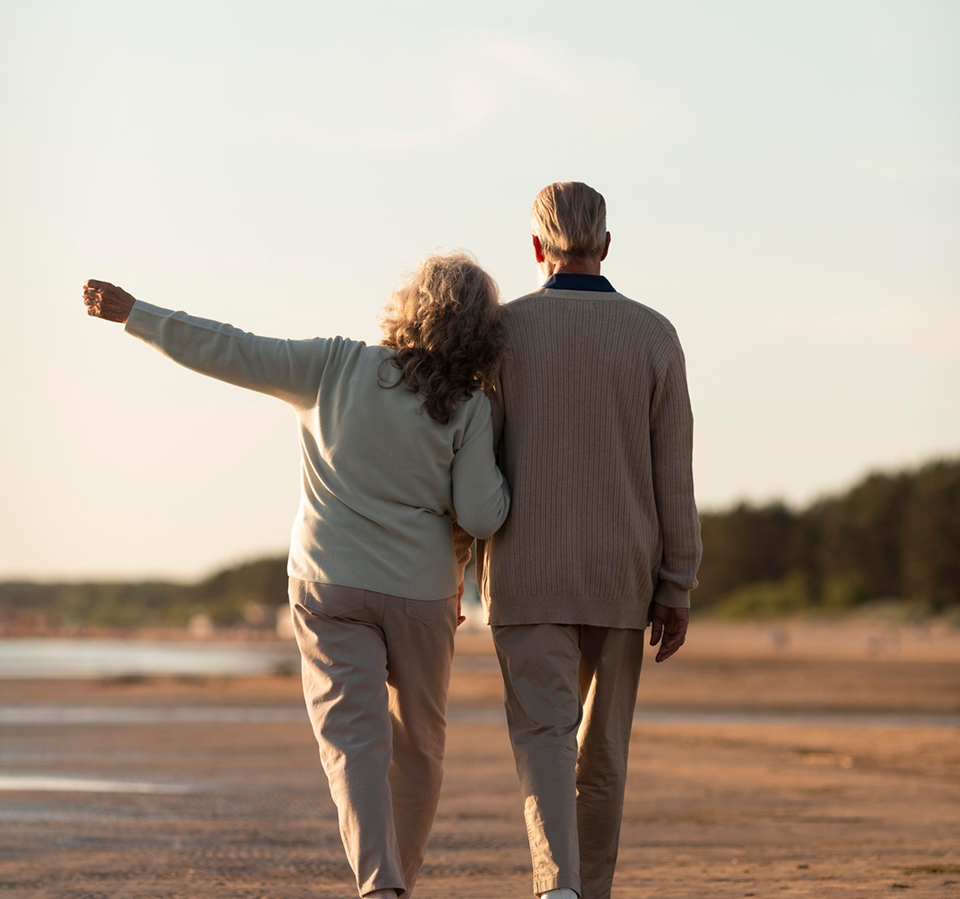Older couple walking together on beach at sunset, woman pointing toward horizon