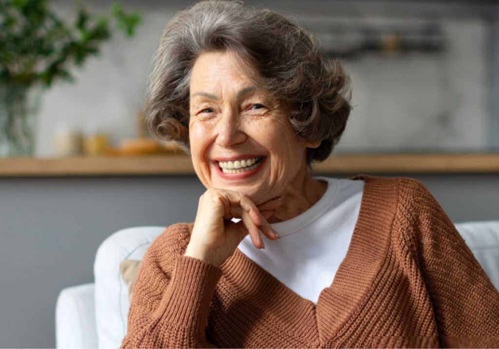 Smiling older woman with gray hair wearing a brown cardigan, resting her head on her hand indoors