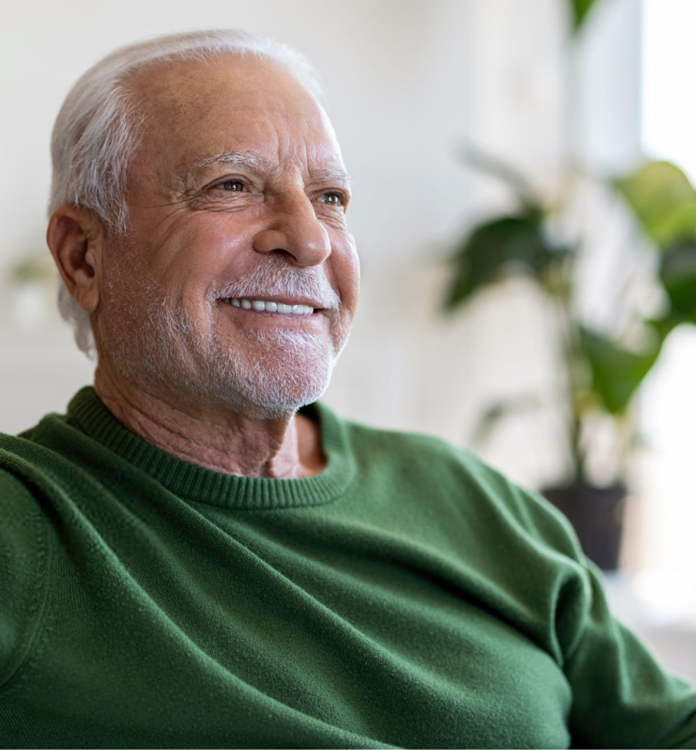 Older man with white hair wearing green sweater, smiling indoors with plants in background