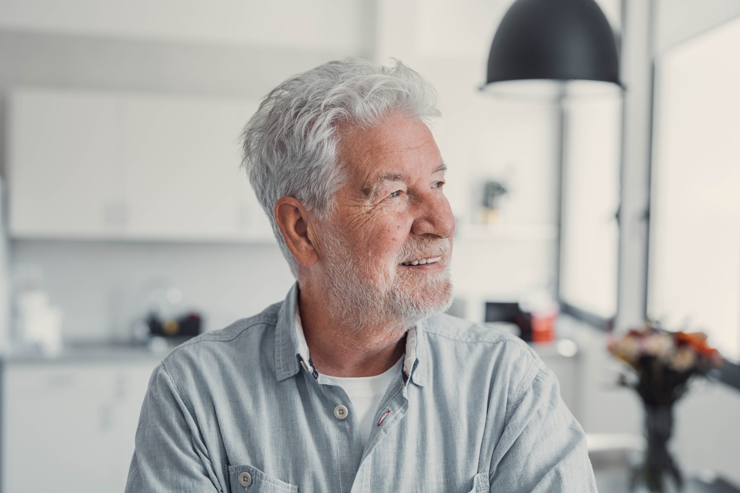 Older man with gray beard in light blue shirt looking thoughtful indoors