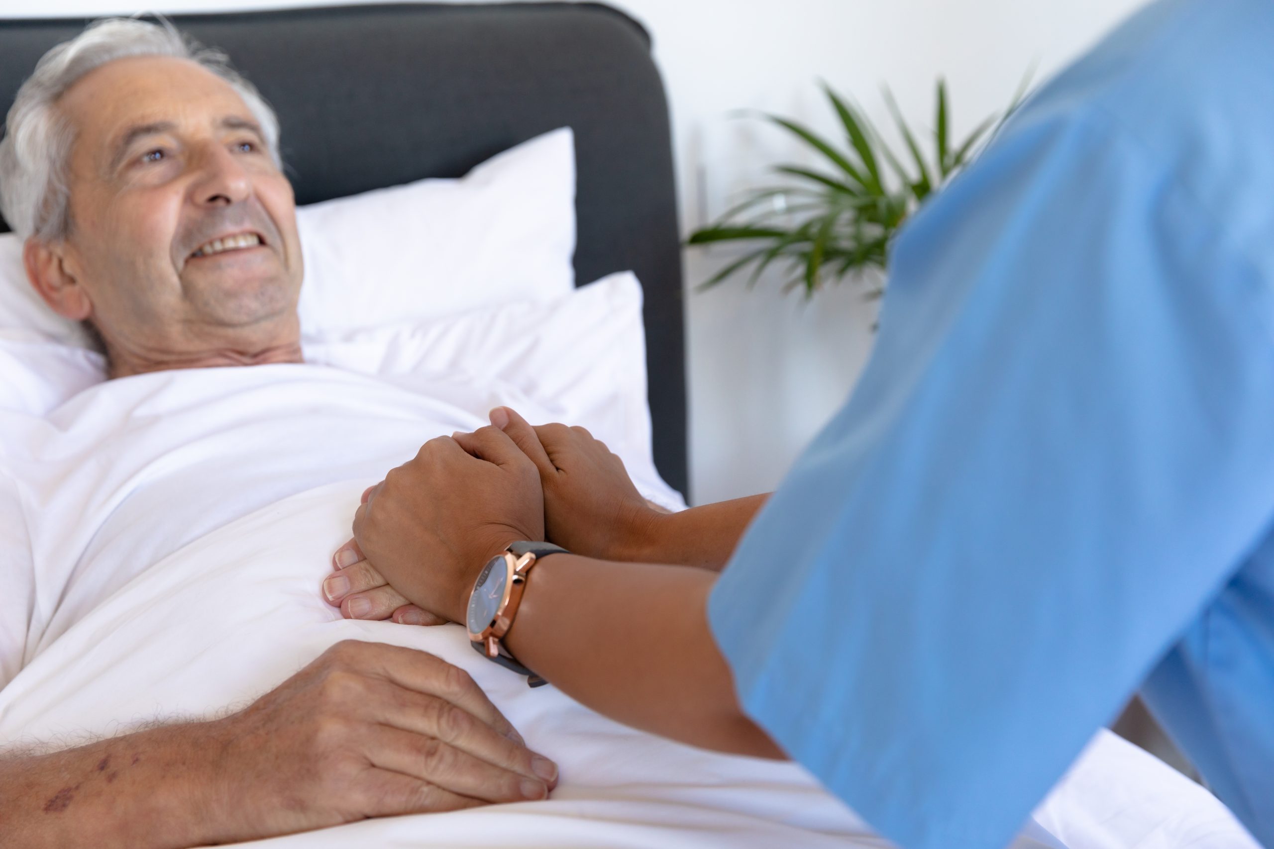 Healthcare worker in blue uniform holding hands with senior patient in hospital bed