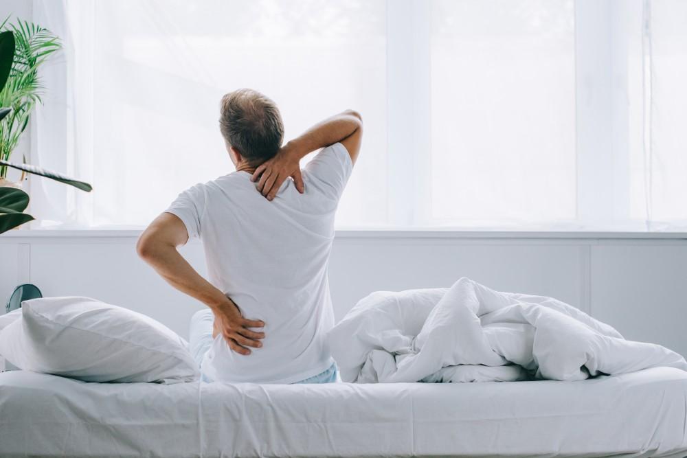 Man sitting on bed holding his lower back and neck in discomfort