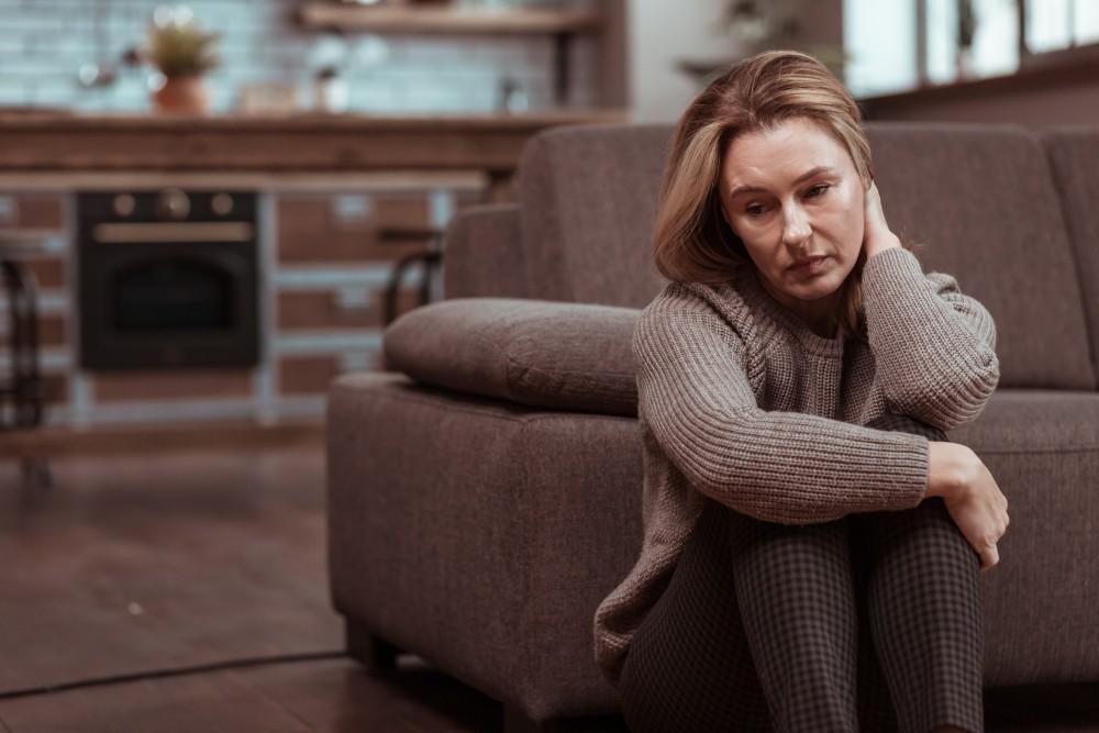 Woman sitting on couch in kitchen, hugging knees with worried expression