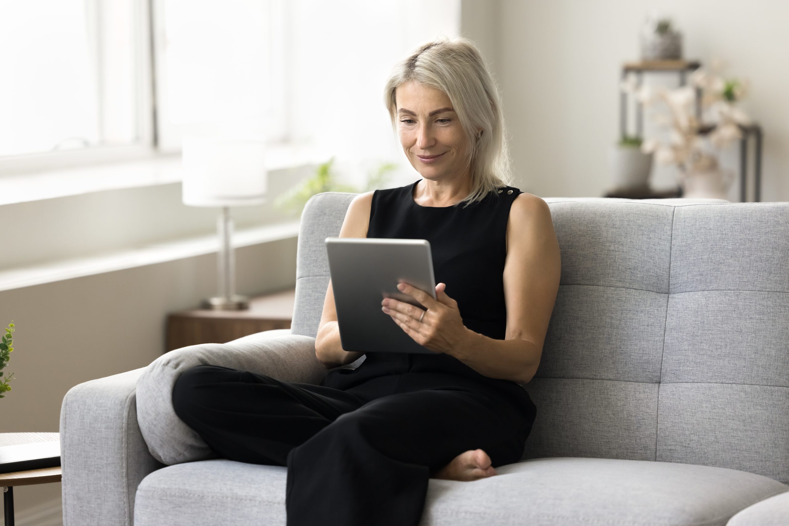 Woman sitting on gray couch looking at tablet device in bright, minimalist living room