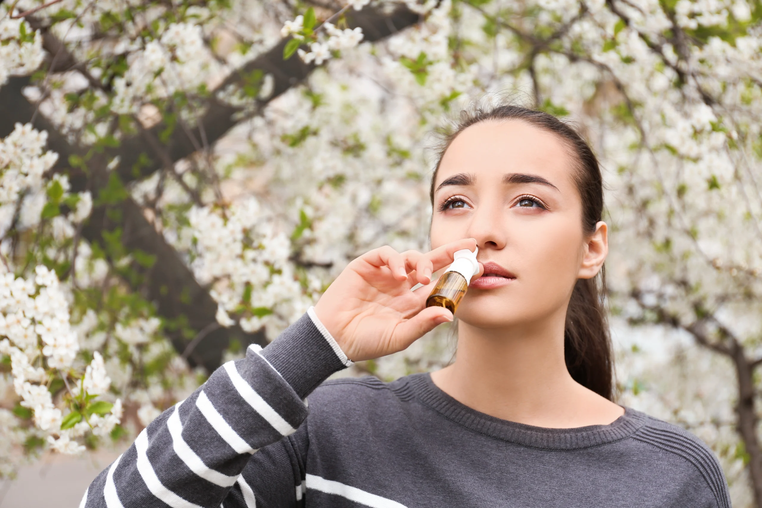 Young woman using a nasal spray outdoors under blossoming trees