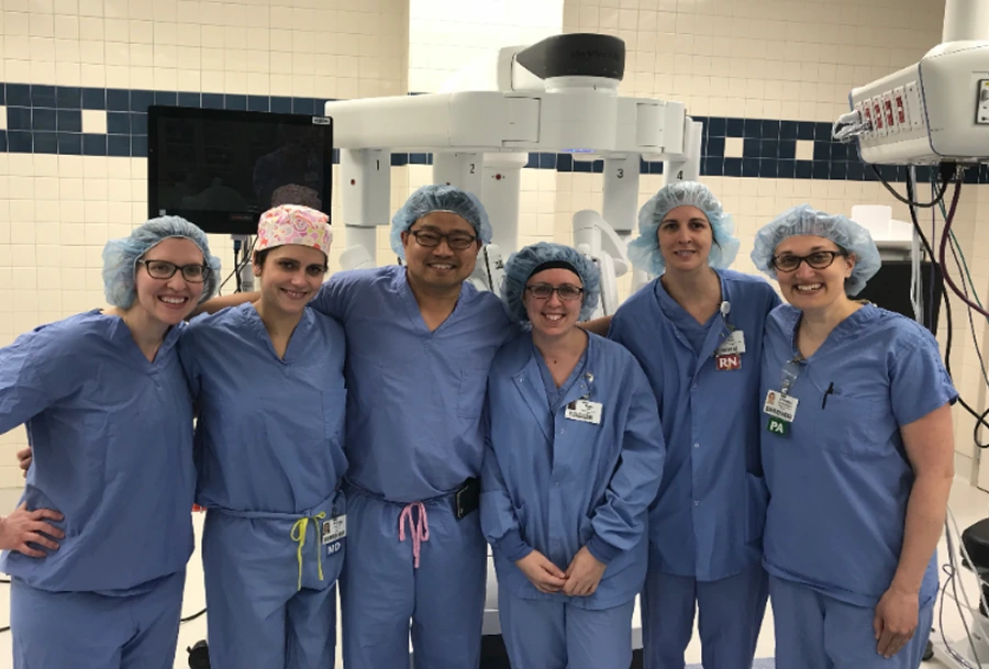 Six surgical team members in blue scrubs and hair covers posing together in operating room