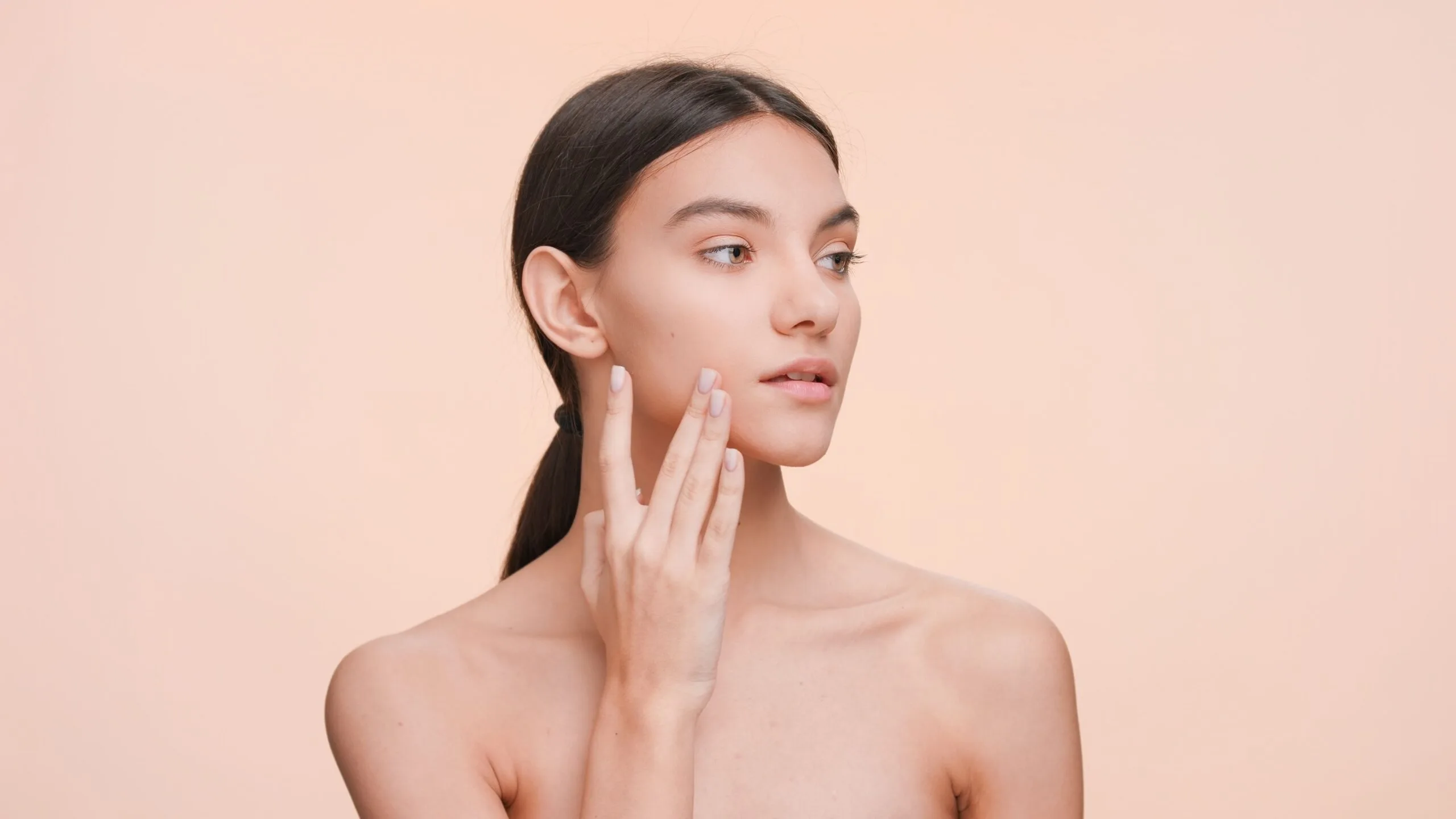 Woman touching her jawline and cheek against peachy background