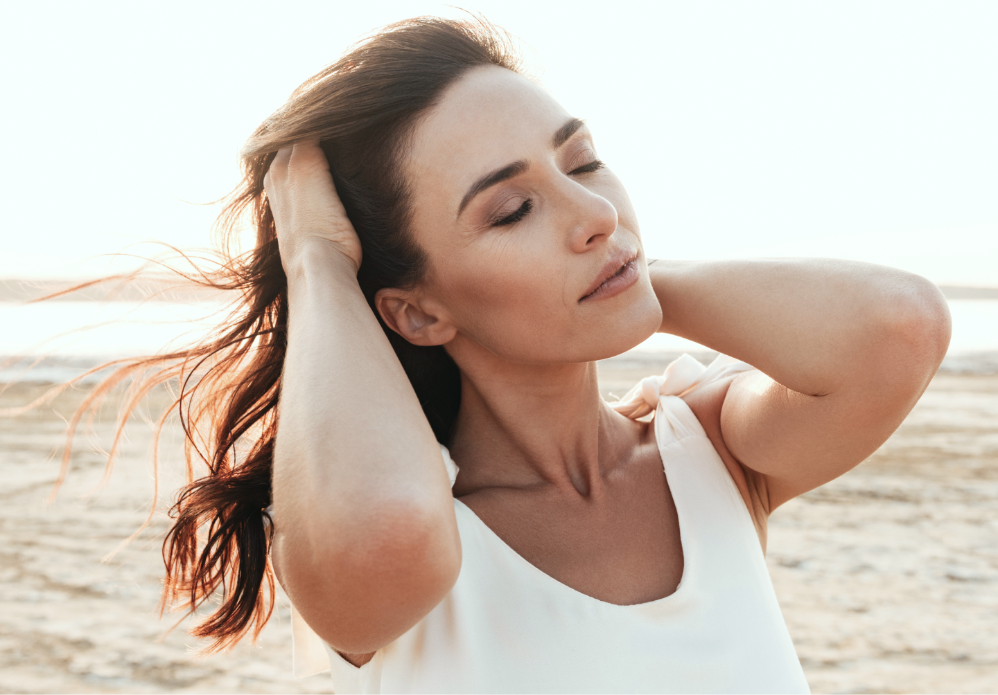 Woman on beach in white tank top with hands behind head, relaxed pose with ocean backdrop