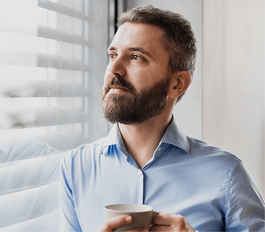 Man with beard holding a cup gazing thoughtfully out a window