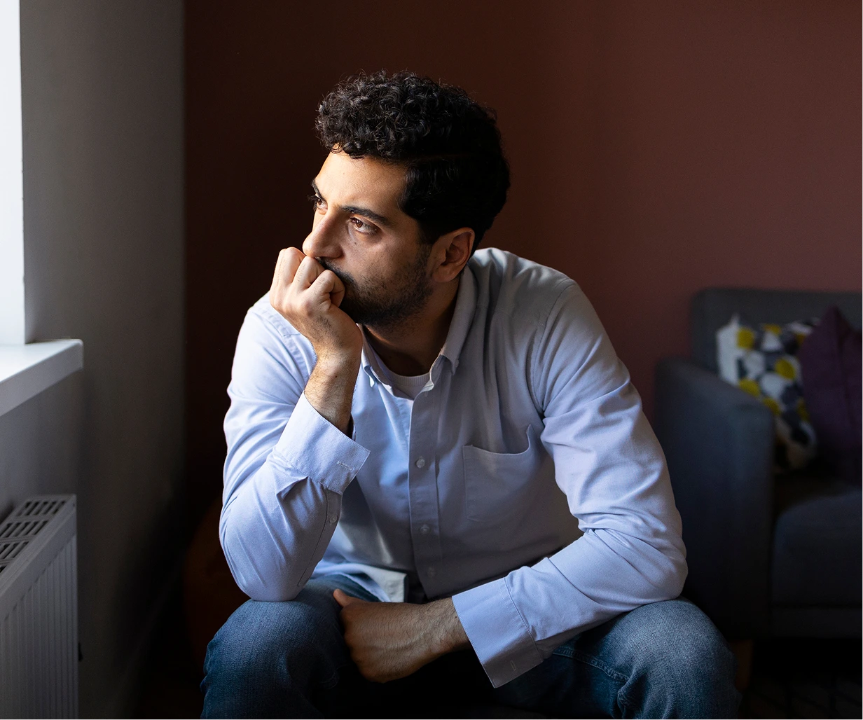 Man in light blue shirt sitting against brown wall, hand to face, displaying anxious expression