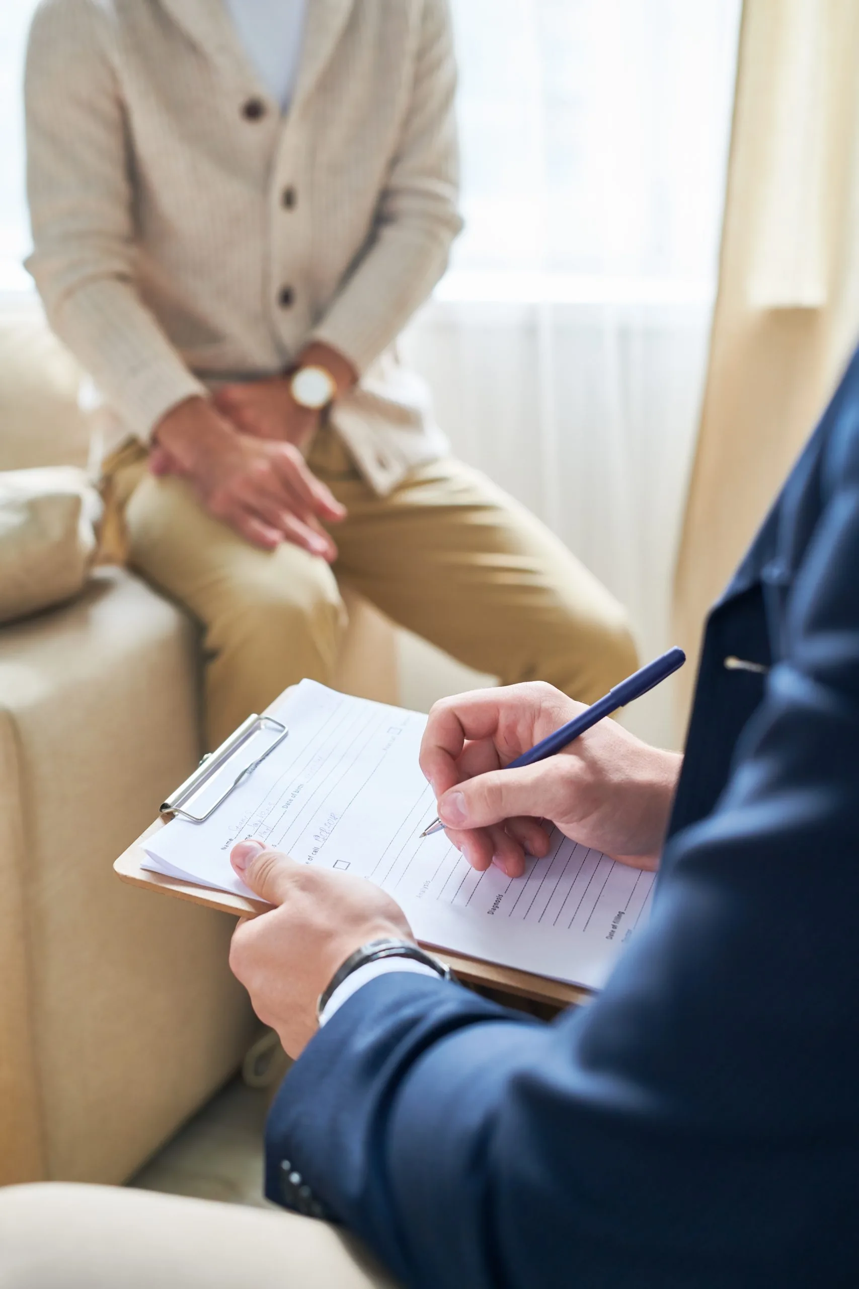 Healthcare provider in dark blazer writing on clipboard while patient sits on couch