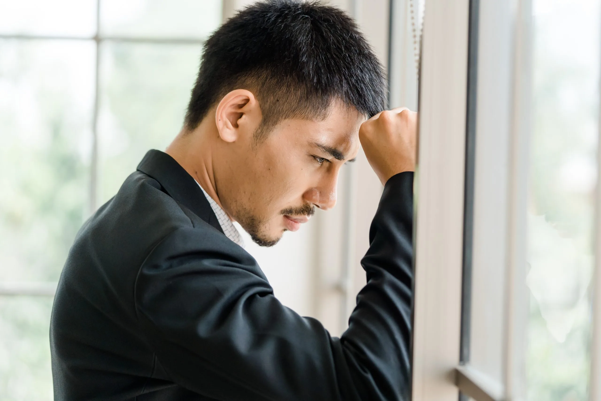 Man in dark jacket leaning against window with hand to head, looking troubled
