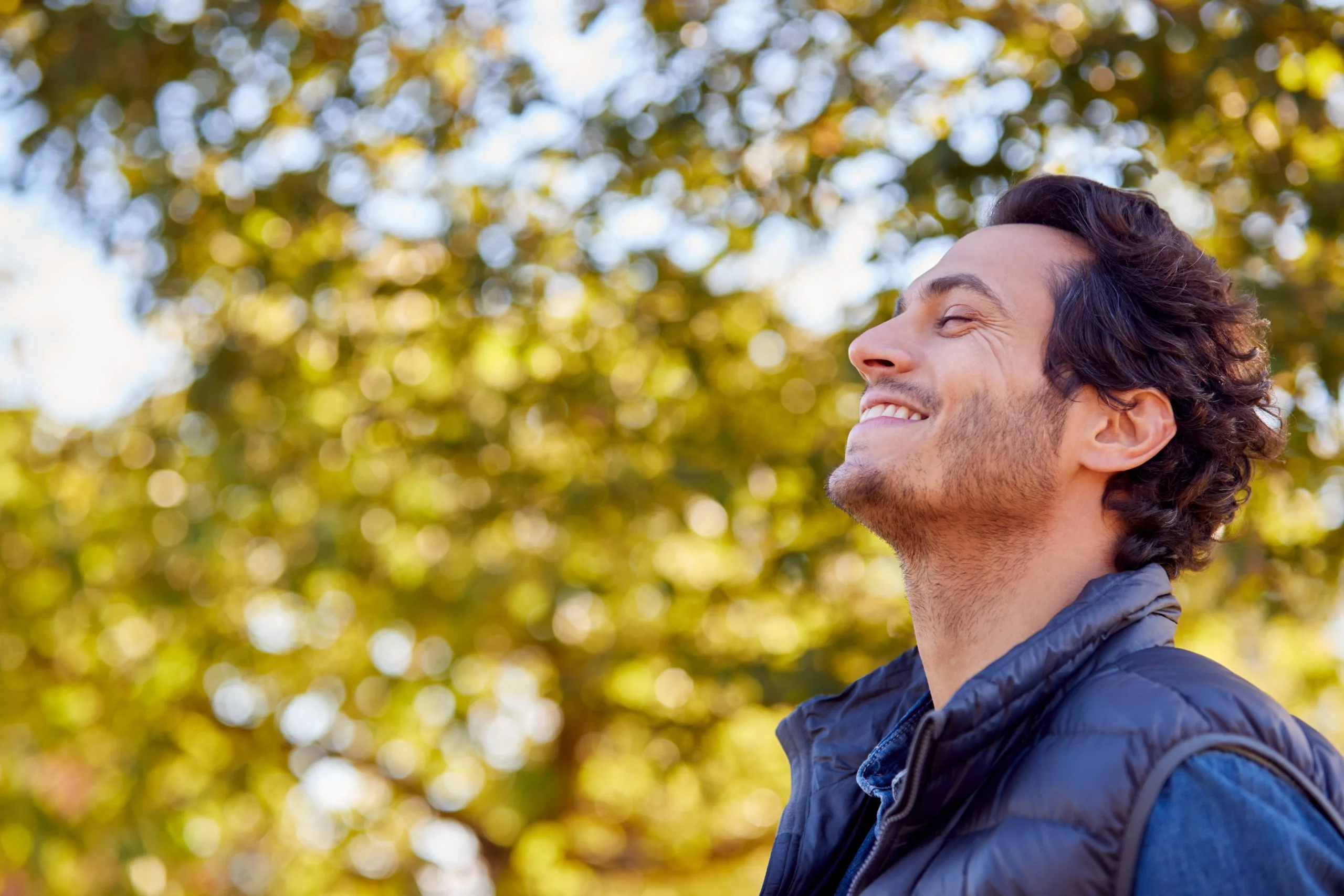 Man in dark blue jacket smiling outdoors with blurred green foliage background