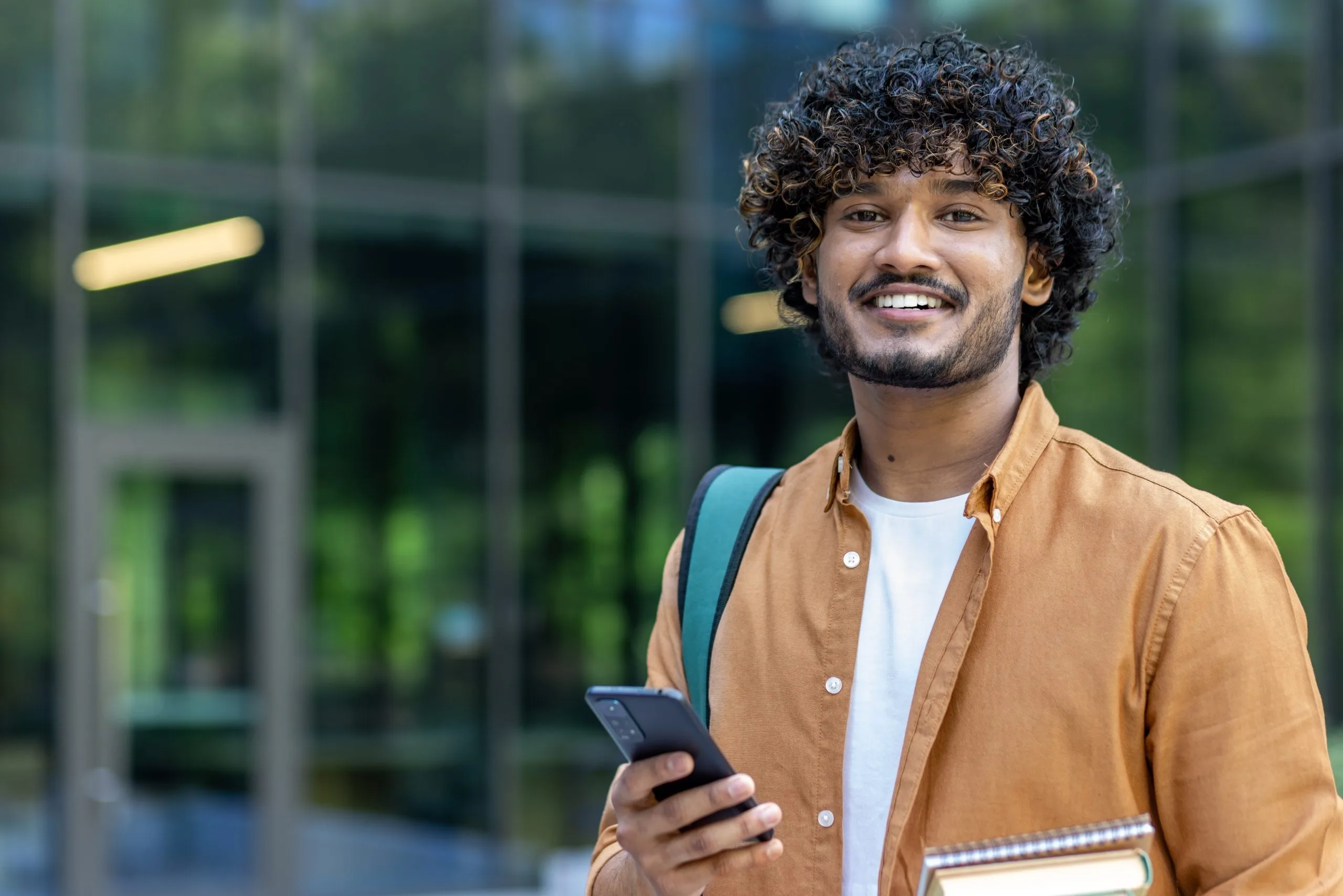 Young man with curly hair holding phone, wearing tan shirt and backpack outdoors
