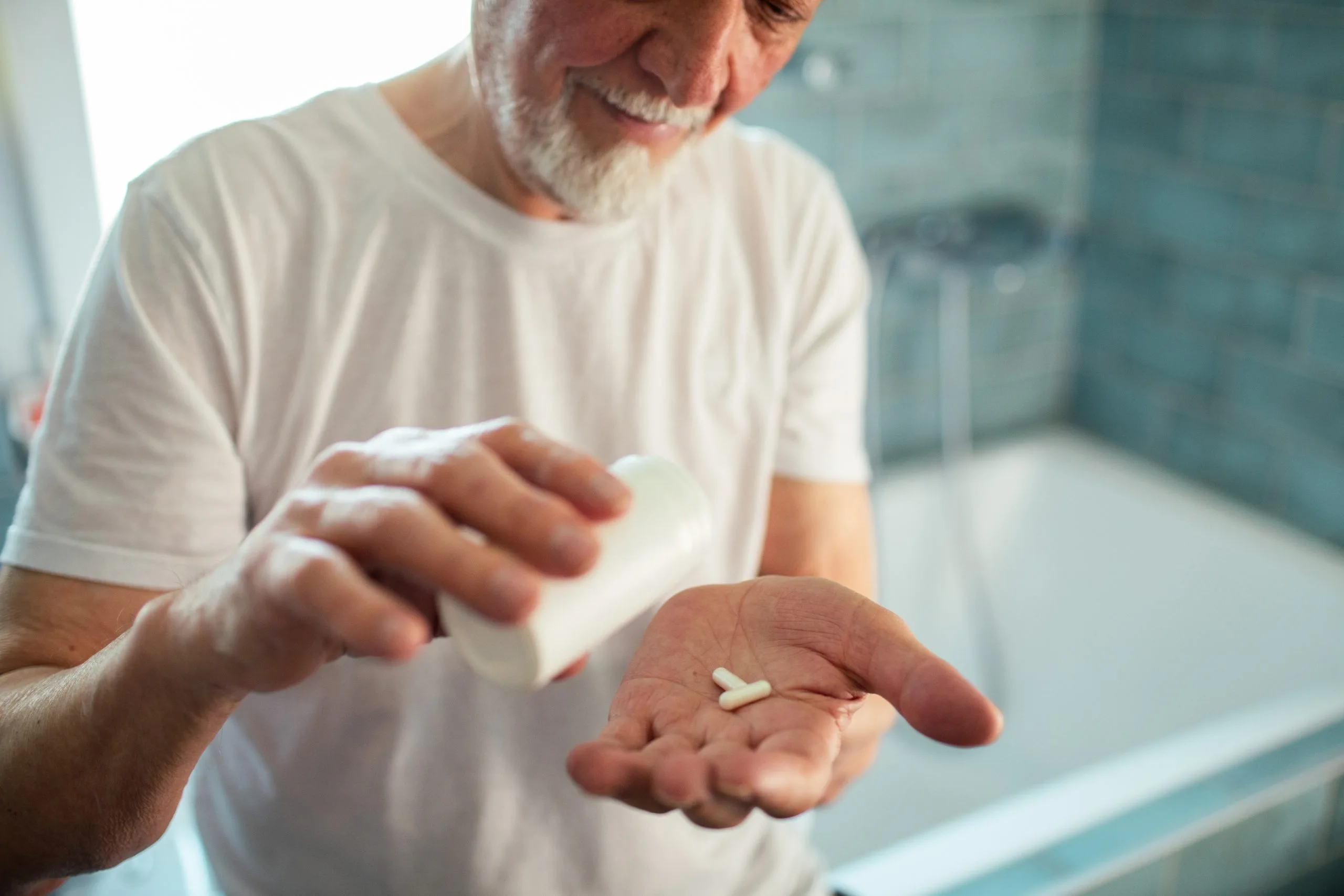 Older man holding white tablets in palm of hand in bathroom setting