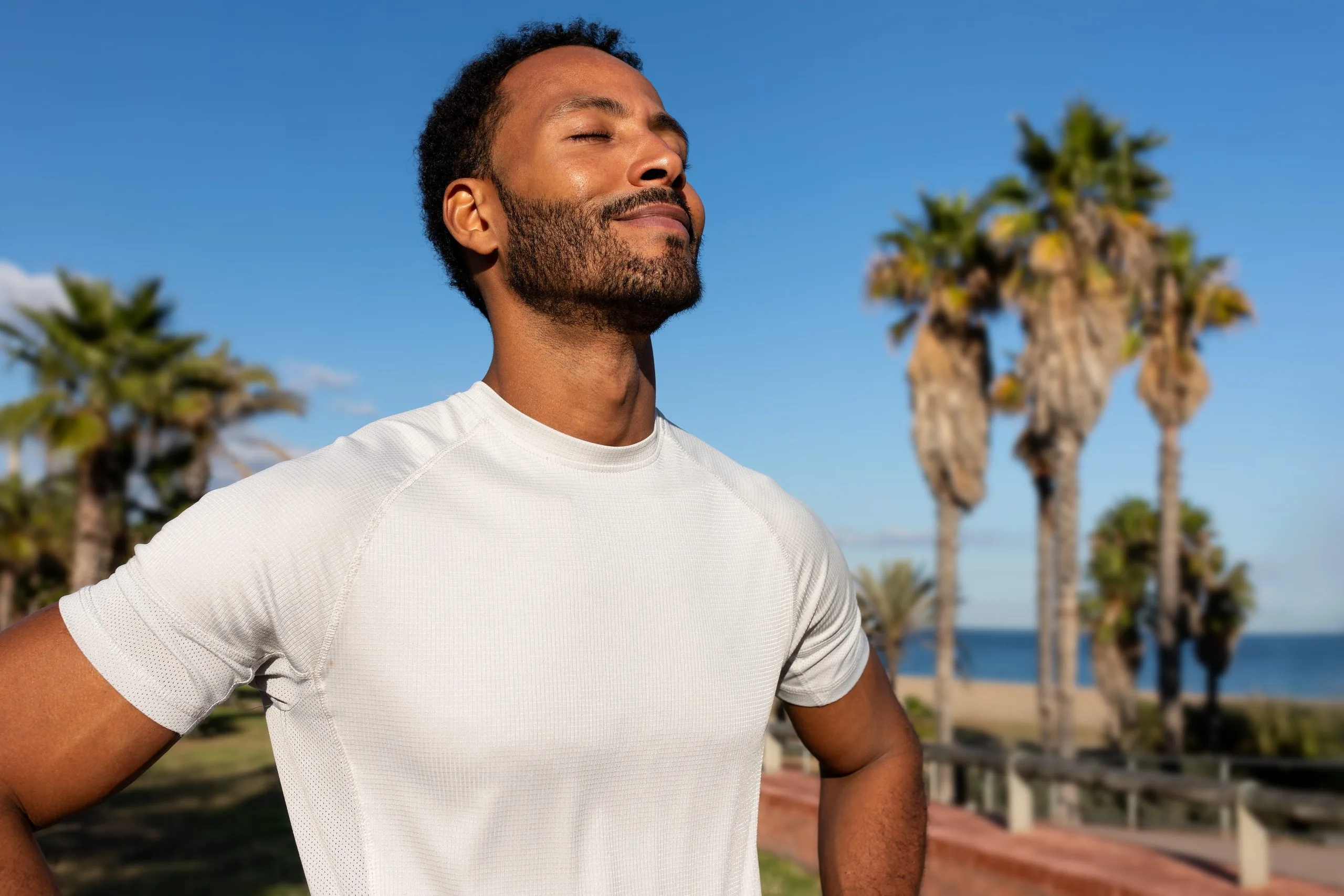 Man in white t-shirt looking upward on a beach with palm trees and ocean